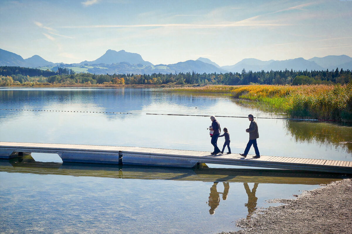 Familie spaziert über einen Holzsteg an einem ruhigen See mit Bergkulisse im Hintergrund.